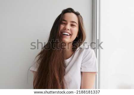 Delighted young lady with long dark hair in t shirt smiling happily and looking at camera, while sitting near window and leaning on white wall Royalty-Free Stock Photo #2188181337