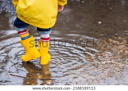 A small child in rainbow socks, yellow rubber boots and a jacket runs through puddles, has fun and plays after the rain. A picture of summer and autumn holidays. Legs close-up.