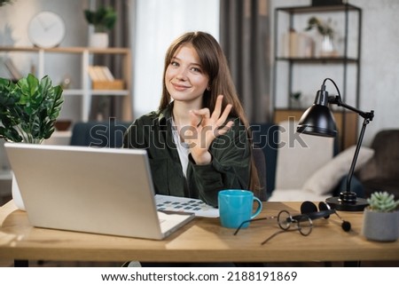Beautiful caucasian woman with long dark hair sitting at desk and typing on laptop. Focused female in dark shirt and jeans using portable computer for work at bright office showing sign ok