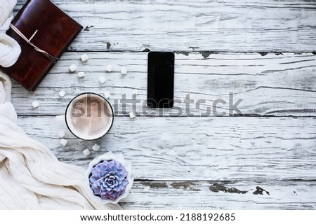 Autumn or winter background shot from top view with hot cocoa, houseplant, cell phone, book, and throw blanket over rustic white wood table. Overhead top view.