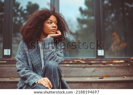 Outdoor autumn portrait of young thoughtful african american woman looking aside sitting on old wooden house stairs, pensive curly girl spending time outside on fall day, thinking about life Royalty-Free Stock Photo #2188194697