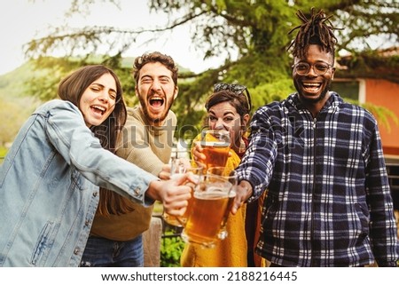 Cheers - multiracial group of happy friends toasting joining beer mugs and laughing looking at the camera standing together outdoors in the farmhouse courtyard - people and alcohol lifestyle concept