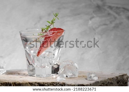 Vodka in shot glass with ice, decorated with strawberry slice and thyme on travertine podium on pink marble stone background. Alcohol cocktail shot with strawberry and vodka, fresh summer shots party