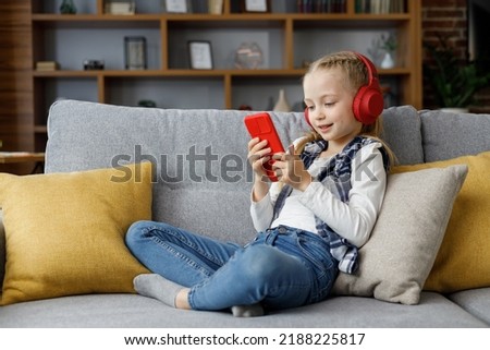 Portrait of cute little cute girl wearing earphones resting at sofa at home. Choosing favorite music or cartoons,texting messages,browsing internet,watching video, playing games on smartphone.