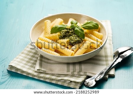 Italian pasta tortiglioni with green basil pesto sauce and fresh basil leaves in white bowl on green checkered kitchen towel on blue wooden background