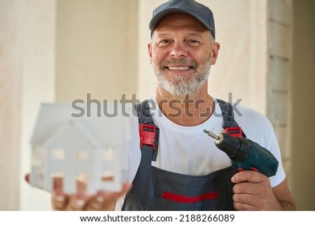 Photo of smiling adult builder holding house model and drill