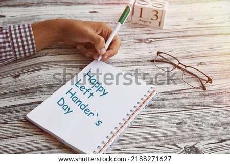 Man uses his left hand to wright in a notebook. Glasess and calendar with 13th of August - Day of Left Handers. Conceptual photo of lefties day. Copy space