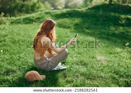A young happy woman sitting in the park on the green grass in nature with her phone in her hands in her casual clothes and chatting via video link, the concept of healthy recreation meditation  Royalty-Free Stock Photo #2188282561