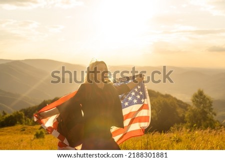 Young happy american woman with long hair holding waving on wind USA national flag on her sholders relaxing outdoors enjoying warm summer day