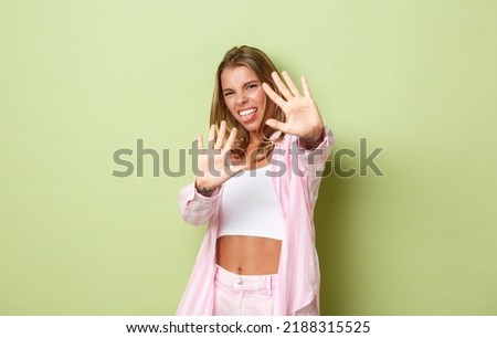 Image of displeased young blond woman defending herself from bright light, raising hands and grimacing bothered, asking to stop taking pictures of her, standing over green background