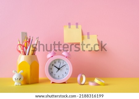 Back to school concept. Photo of school supplies pink alarm clock adhesive tape stand for pens bunny shaped sharpener and sticky note paper attached to pink wall