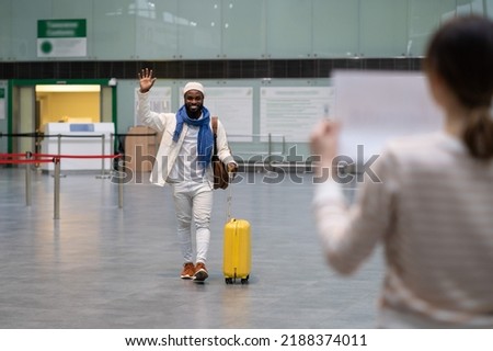 Young happy excited african american man with luggage waving with hand to female friend waiting for him in airport, joyful guy arriving in terminal, moving overseas, travelling by plane Royalty-Free Stock Photo #2188374011