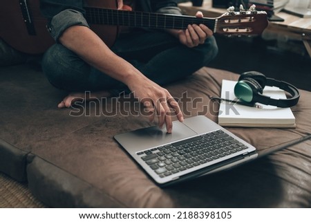 young man relax and playing guitar while sitting on sofa bed in living room at home. Music create melody song, lyrics on laptop and practice concept. Royalty-Free Stock Photo #2188398105