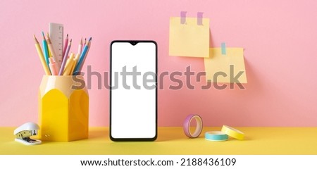Back to school concept. Photo of school supplies on yellow desk smartphone pencil holder adhesive tape stapler and sticky note paper attached to pink wall with blank space