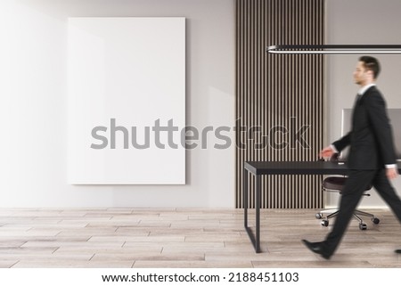 Side view of young european businessman walking in office interior with furniture, equipment, wooden flooring and empty white mock up poster on concrete wall. CEO, executive and worker concept