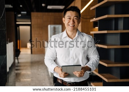 Portrait of smiling cheerful positive asian businessman in white shirt with tablet looking directly in camera indoor Royalty-Free Stock Photo #2188455101