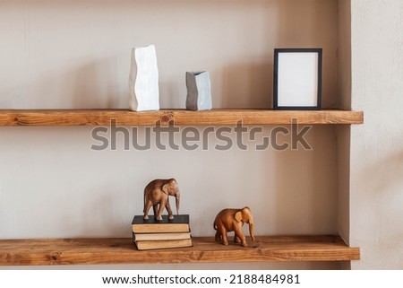 Grey and white colored vases, wooden figures of elephants, blank black photoframe and stack of books on wooden shelf against  grey wall. Home decor.
