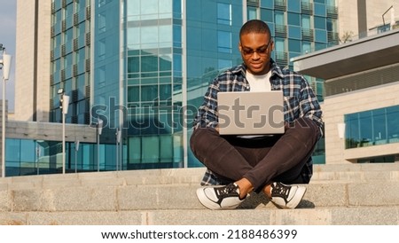 Concentrated young business man african american male freelancer guy student in sunglasses sitting city background company building with laptop working typing learning online using wireless computer