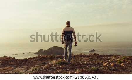Travel photographer walking towards the cliff to take a panoramic landscape photo at sunset. Travel photographer from behind with camera in hand looking at an island on the horizon