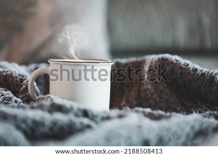 White enamel cup of hot steaming coffee sitting on a cozy bed surrounded by throw pillows and knit blankets. Selective focus on mug with extreme blurred foreground and background.