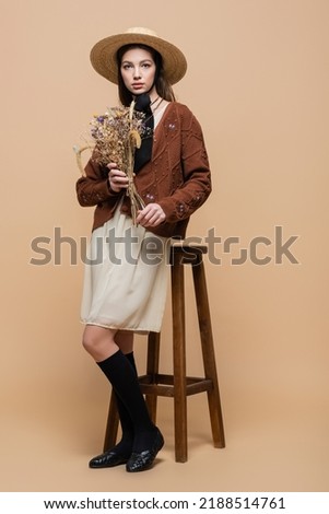Full length of trendy woman in straw hat holding flowers near chair on beige background Royalty-Free Stock Photo #2188514761
