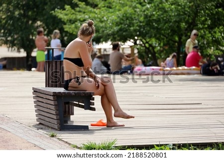 Woman wearing black swimsuit sitting on wooden bench and talking on mobile phone. Hot weather in city, leisure and tanning in summer park Royalty-Free Stock Photo #2188520801