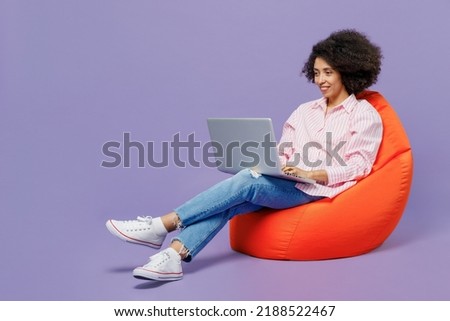 Full body young woman of African American ethnicity 20s wear pink striped shirt sit in bag chair hold use work on laptop pc computer isolated on plain pastel light purple background studio portrait