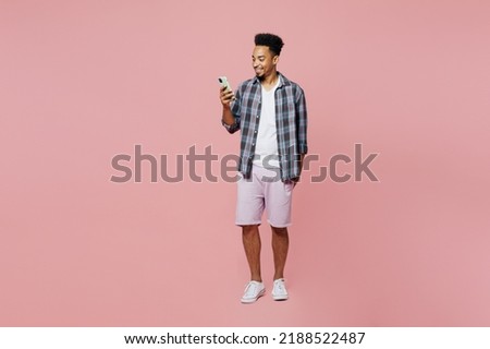Full body young smiling man of African American ethnicity he wear blue shirt hold in hand use mobile cell phone isolated on plain pastel light pink background studio portrait. People lifestyle concept