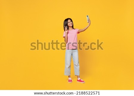 Full body little kid girl of African American ethnicity 12-13 years old in pink t-shirt do selfie shot on mobile cell phone show v-sign isolated on plain yellow background. Childhood lifestyle concept