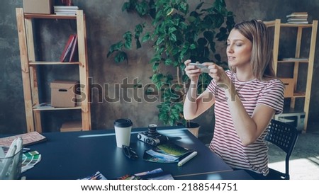 Young woman is arranging pictures, take-out coffee, sun glasses, professional camera and marker on table and shooting flat lay with smartphone, then checking photos by touching screen.
