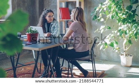 Young enterpreneurs are duscussing advertising strategy while sitting together at table in modern office. Attractive women are talking and gesturing, one of them is making notes. Royalty-Free Stock Photo #2188544771