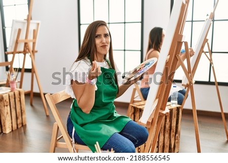 Young hispanic artist women painting on canvas at art studio doing money gesture with hands, asking for salary payment, millionaire business 