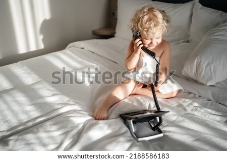blonde child with curly blond hair rings the phone in a hotel room while sitting on a bed in a towel. In the morning light