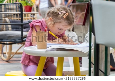 Street portrait of a little girl drawing with colored pencils at a table in an outdoor cafe on a blurry background of the old town.