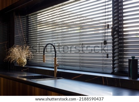 Wooden blinds black color closeup on the window. Bamboo slats 50mm wide. Venetian wood blinds in the kitchen. Black tapes. Sink with copper faucet near the window. Round vase is on the windowsill. Royalty-Free Stock Photo #2188590437