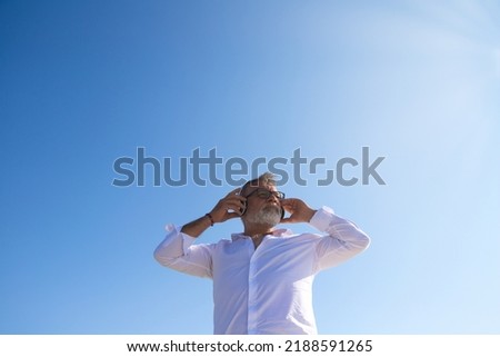 handsome mature man with beard and grey hair is in the park while listening to music with his headphones. Picture taken from below and the blue sky in the background. Senior concept, travel and music.