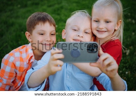 cute funny children shooting selfie with smartphone. Kids in park sitting on grass having fun together. Image with selective focus