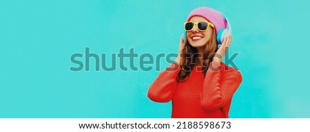Portrait of happy smiling young woman in wireless headphones listening to music wearing red knitted sweater, pink hat on blue background