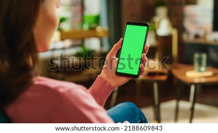 Feminine Hand Tapping on Display and Scrolling Feed on Smartphone with Green Screen Mock Up Display. Female Resting at Home, Checking Social Media on Mobile Device. Close Up Over the Shoulder Photo.