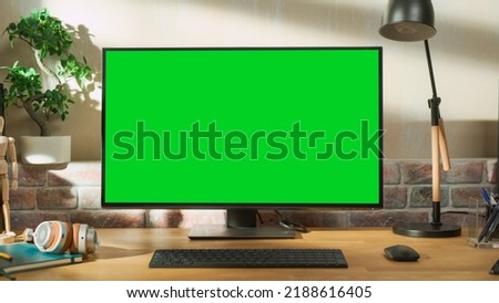 Desktop Personal Computer Monitor Standing on a Table with a Green Screen Chromakey Mock Up Display. Cozy Empty Loft Apartment with a Lamp, Notebooks and Headphones on the Table.