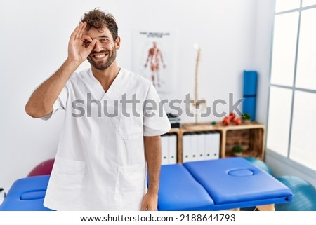 Young handsome physiotherapist man working at pain recovery clinic doing ok gesture with hand smiling, eye looking through fingers with happy face. 