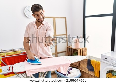 Young man with beard ironing clothes at home smiling cheerful offering palm hand giving assistance and acceptance. 