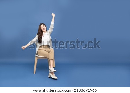 Attractive beautiful Asian woman sitting on white modern chair and hands up raised arms from happiness isolated on blue background, Excited businesswoman winner success concept