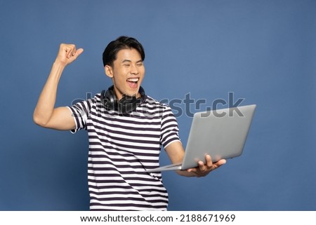 Happy young Asian pro gamer with headphones holding computer laptop and win in online video game isolated over blue background, Cyber sportsman playing tournaments concept