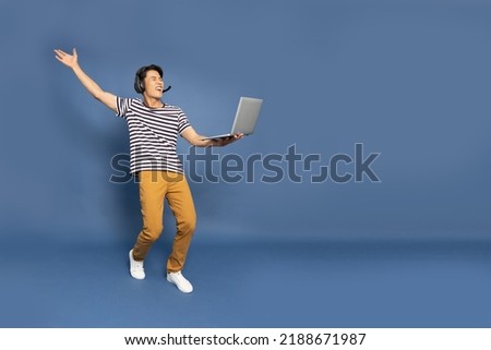 Happy young Asian pro gamer with headphones holding computer laptop and win in online video game isolated over blue background, Full body composition, Cyber sportsman playing tournaments concept
