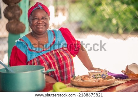Portrait of an adult woman standing in her kitchen looking at a camera. Proud African American woman in colorful Garifuna dress. Royalty-Free Stock Photo #2188675821