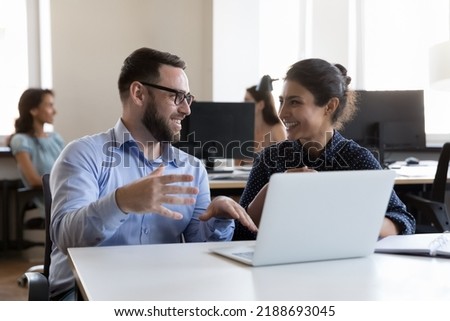 Millennial Indian woman and Caucasian man colleagues smile while working on project sit in co-working workspace with laptop. Teamwork, friendly relations at workplace, tech, creative workflow
