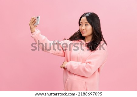 Cheerful smiling Asian young lady in pink hoodie sweatshirt doing selfie video call distance communication posing isolated on pink studio background. Good offer. Gadget addiction Social Media