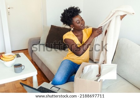 Beautiful young curly hair satisfied happy African woman shopaholic customer sit on sofa unpack parcel delivery box look at clothes take out beige sweater, online shopping shipment concept.