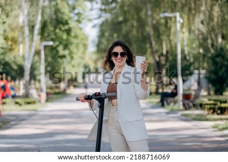 Beautiful young woman in white suit and sunglasses standing with her electric scooter in city parkland and drinking coffee